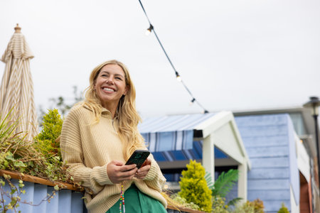 Wide-view shot of a mid adult woman standing in a city centre street in Newcastle, England. She is wearing casual clothing, smiling and looking away while using a smartphone.の写真素材