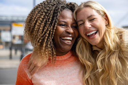 Front-view shot of two mid adult female friends standing in a city street in Newcastle, England. They are wearing casual clothing and sharing a laugh with their heads together.の写真素材