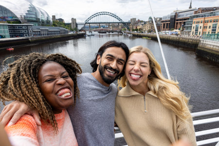 Front-view selfie shot of three mid adult friends crossing a bridge in Newcastle, England. They are smiling and looking at the camera while taking a silly group selfie.の写真素材