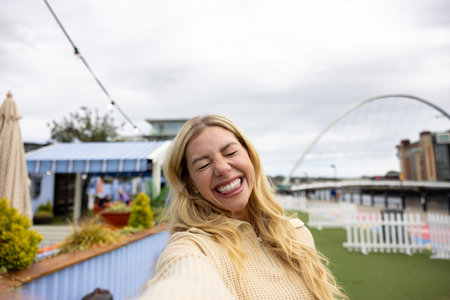 Front-view of a mid adult woman standing on a bridge. She is wearing casual clothing, smiling with her eyes closed, while taking a selfie on a smartphone.の写真素材