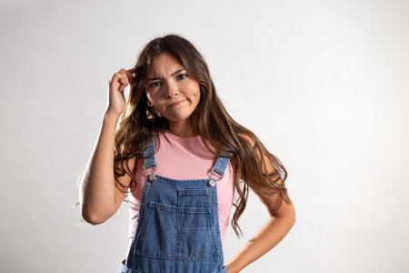 Waist-up shot of a young adult female looking at the camera, one hand on hip and the other scratching her head. She has long brunette hair and is wearing denim dungarees.の写真素材