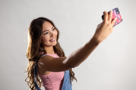 Waist-up shot of a young adult female holding a mobile phone, smiling as she captures a selfie. She has long brunette hair and is wearing denim dungarees with a pink top.の写真素材