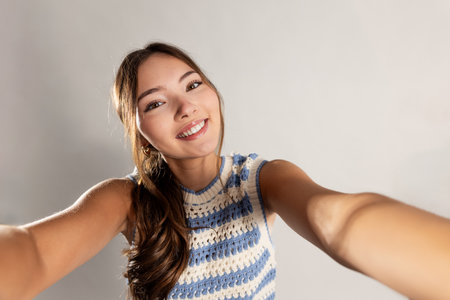Point-of-view shot of a young adult female reaching towards the camera as though she is capturing a selfie. She has long brunette hair and is wearing a knitted tank top.の写真素材
