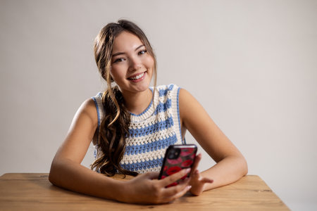 Waist-up portrait of a young adult female sitting at a table, smiling. She is leaning on the table, holding a mobile phone. She has long brunette hair and is wearing a knitted top.の写真素材