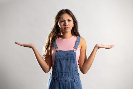 Waist-up shot of a young adult female looking at the camera with a confused expression, both hands extended to the sides. She has long brunette hair and is wearing denim dungarees.の写真素材