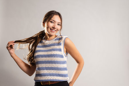 Waist-up shot of a young adult female smiling at the camera with one hand on her hip and the other holding her ponytail. She has long brunette hair, wearing a knitted tank top.の写真素材