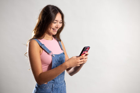 Waist-up shot of a young adult female holding a mobile phone, looking down with a positive expression. She has long brunette hair and is wearing denim dungarees with a pink top.の写真素材