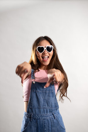 Waist-up shot of a young adult female pointing towards the camera with a positive expression. She has long brunette hair, wearing heart-shaped sunglasses and denim dungarees.の写真素材