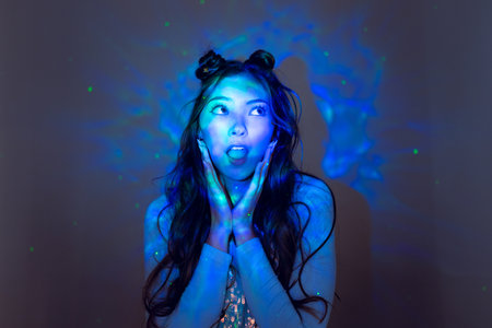 Waist-up shot of a young adult female, looking off-frame with a surprised expression. She has long brunette hair with space buns. There is colourful neon light shining on her.の写真素材