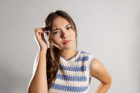 Studio portrait of a young adult female looking at the camera with a puzzled expression, scratching her head. She has long brunette hair and is wearing a knitted tank top.の写真素材