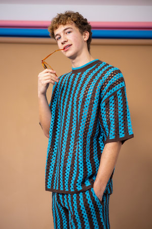 Three-quarter-length studio shot of a young adult male looking directly at the camera with a neutral expression, standing with one hand in his pocket and the other holding a pair of sunglasses which he holds between his teeth. He has brown curly hair and is wearing a blue and brown striped crochet coordinated outfit.の写真素材