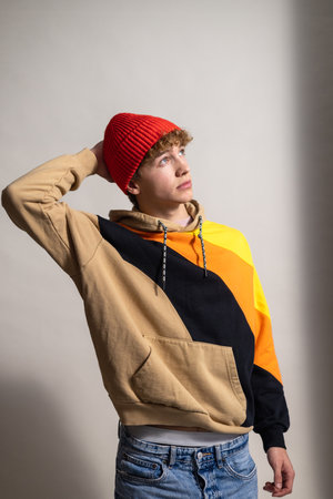 Waist-up studio shot of a young adult male looking off-frame with a neutral expression. He is wearing a knitted hat and hooded sweatshirt, one hand resting behind his head. His brown curly hair is visible beneath the hat.の写真素材