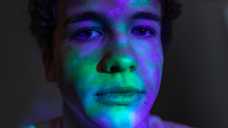 Extreme close-up creative studio headshot of a young adult male looking directly at the camera with a serious expression. There are neon coloured lights shining on his face creating various patterns.の写真素材