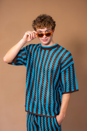 Three-quarter-length studio shot of a young adult male looking directly at the camera with a neutral expression. He is wearing sunglasses with a blue and brown striped crochet coordinatied outfit, one hand in his pocket while he holds his sunglasses below his eyes with the other. He is standing against a light brown background.の写真素材