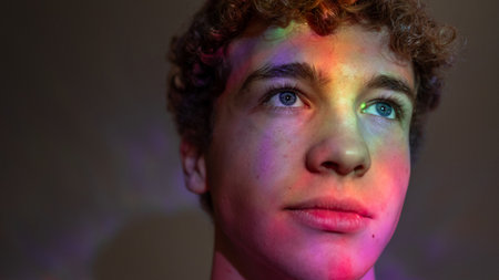 Close-up creative studio headshot of a young adult male with brown curly hair looking off-frame with a neutral expression. There are neon coloured lights shining on his face creating various patterns.の写真素材