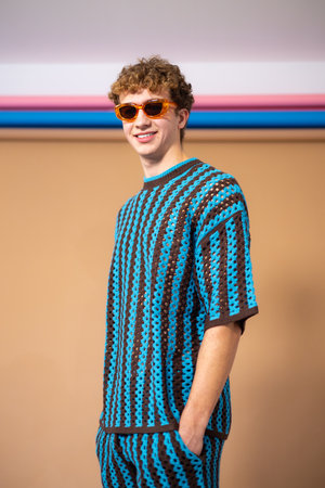 Three-quarter-length studio shot of a young adult male smiling directly at the camera, wearing sunglasses with his hands in his pockets. He has brown curly hair and is wearing a blue and brown striped crochet coordinated outfit.の写真素材