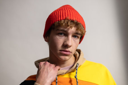 Close-up studio headshot of a young adult male looking directly at the camera with a serious expression. He is wearing a knitted hat and hooded sweatshirt which he is holding the collar of with one hand. His brown curly hair is visible beneath the hat.の写真素材