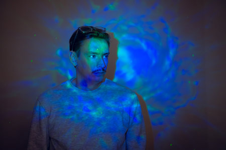 Creative studio shot of a young adult male with a moustache looking off-frame with a neutral expression. There are neon coloured lights shining on him in patterns.の写真素材