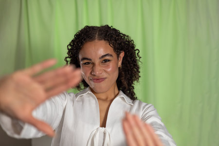 Studio shot of a young adult female with curly hair, making a scrunched face towards the camera. She is wearing a white shirt, reaching both hands towards the camera.の写真素材