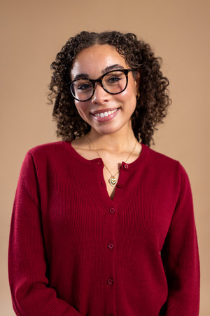 Waist-up studio shot of a young adult female smiling at the camera. She has curly hair and is wearing a smart red cardigan, standing against a light brown background.の写真素材