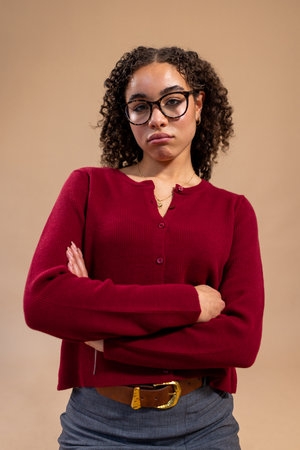 Waist-up studio shot of a young adult female standing with her arms folded, frowning at the camera. She is wearing glasses and smart-casual clothing, standing against a light brown background.の写真素材