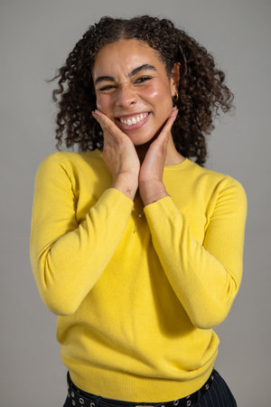 Waist-up studio shot of a young adult female smiling at the camera with her hands rasied to her face. She has curly hair and is wearing a yellow jumper, standing against a grey background.の写真素材