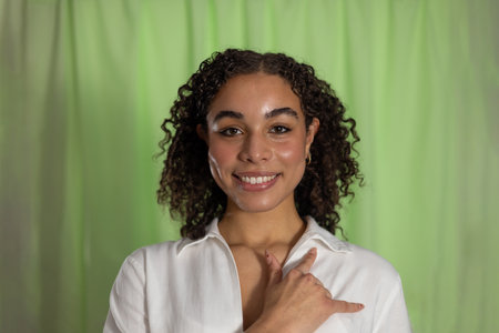 Studio headshot of a smiling young adult female with curly hair. She is looking at the camera, wearing a white shirt and gesturing with one hand in sign language.の写真素材