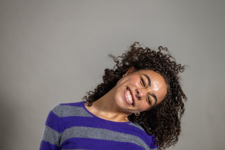Studio shot of a young adult female with curly hair, smiling and making a scrunched face towards the camera with her head tipped to one side. She is wearing a striped jumper, standing against a grey backgorund.の写真素材