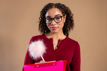 Waist-up studio shot of a young adult female standing holding a clipboard, smiling at the camera. She is wearing glasses and smart-casual clothing.の写真素材