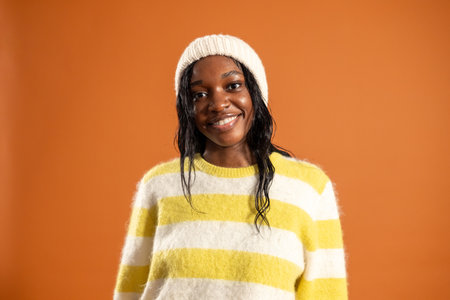 Waist-up studio portrait of a young adult female smiling at the camera. She is wearing casual clothing and a woollen hat, standing against an orange studio background.の写真素材
