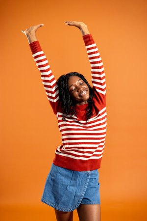 Studio portrait of a young adult female smiling with her eyes closed and arms raised above her head. She is wearing a striped jumper, standing against an orange studio background.の写真素材