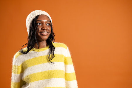 Waist-up studio portrait of a young adult female smiling away from the camera. She is wearing casual clothing and a woollen hat, standing against an orange studio background.の写真素材