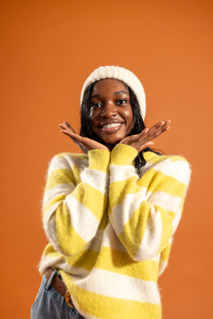 tudio portrait of a young adult female smiling at the camera, hands cupping her face. She is wearing casual clothing and a woollen hat, standing against an orange background.の写真素材