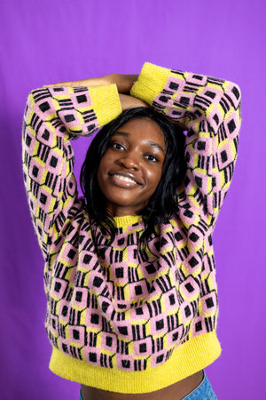 Studio portrait of a young adult female smiling at the camera, both arms resting on her head. She is wearing a funky patterned jumper, standing against a purple studio background.の写真素材