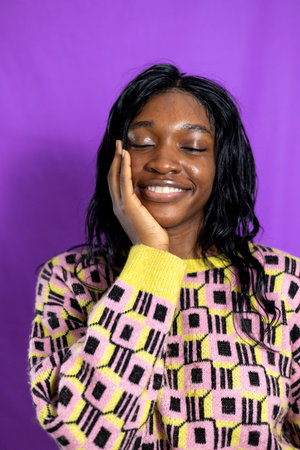 Studio portrait of a young adult female smiling with her eyes closed and one hand cupping her cheek. She is wearing a patterned jumper, standing against a purple studio background.の写真素材