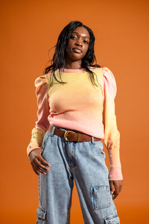 Studio portrait of a young adult female looking at the camera with a serious expression. She is wearing casual clothing, standing against an orange studio background.の写真素材
