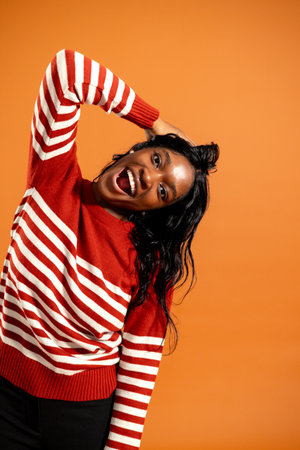 Studio portrait of a young adult female smiling with her hand on her head as it is tipped to one side. She is wearing a striped jumper, standing against an orange background.の写真素材