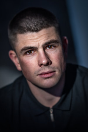 Close-up high contrast studio headshot of a young adult male looking at the camera with a serious expression. He has short dark hair and is wearing a dark coloured top.の写真素材