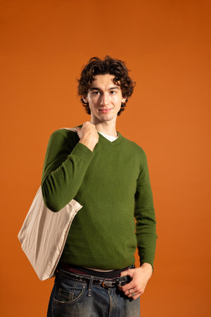 Studio shot of a young adult male standing with a tote bag over one shoulder, looking at the camera. He has curly hair and is wearing casual clothing, against an orange background.の写真素材