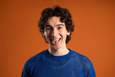 Studio shot of a young adult male smiling and sticking his tongue out at the camera. He has dark curly hair and is wearing casual clothing, standing against an orange background.の写真素材