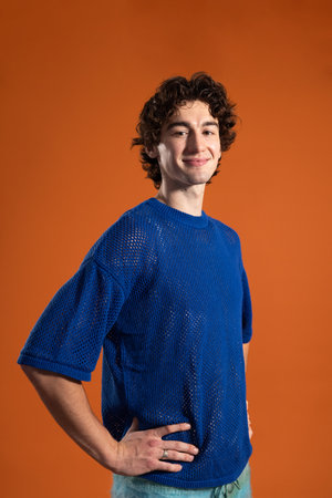 Waist-up studio shot of a young adult male smiling at the camera with hands on hips. He has dark curly hair and is wearing casual clothing, standing against an orange background.の写真素材