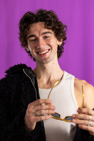 Waist-up studio shot of a young adult male wearing a fluffy jacket, looking at the camera. He has curly hair and is holding sunglasses, standing against a purple background.の写真素材