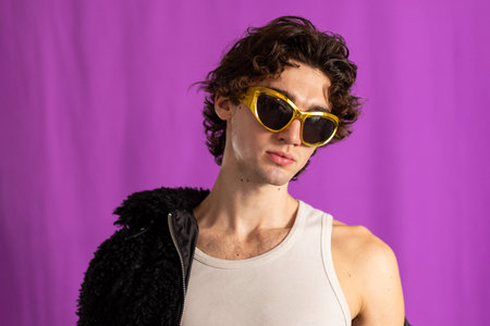 Studio shot of a young adult male with curly hair wearing a fluffy jacket and gold sunglasses, looking away from the camera. His jacket is off one shoulder, the background purple.の写真素材