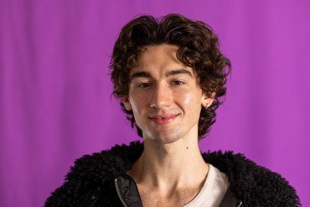 Studio headshot of a young adult male smiling directly at the camera. He has dark curly hair and is wearing a black fluffy jacket, standing against a purple background.の写真素材
