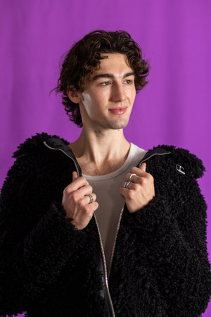 Studio shot of a young adult male wearing a fluffy jacket, looking away from the camera. He has curly hair, holding the edges of his jacket, standing against a purple background.の写真素材