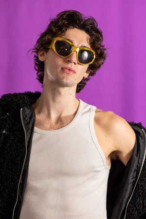 Studio shot of a young adult male with curly hair wearing a fluffy jacket and gold sunglasses, looking away from the camera. His jacket is off one shoulder, the background purple.の写真素材
