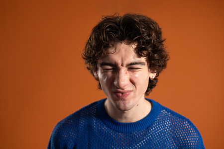 Studio headshot of a young adult male making a scrunched face to the camera. He has dark curly hair and is wearing casual clothing, standing against an orange background.の写真素材