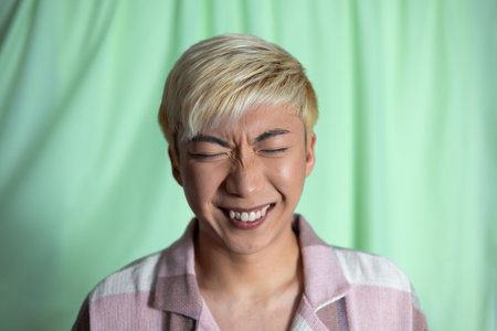 Studio headshot of a young adult male smiling with eyes closed and nose scrunched. He has bleached...