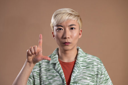 Studio shot of a young adult male gesturing to the camera, making an 'L' shape with one hand. He has bleached hair and is wearing a knitted shirt, against a neutral background.の写真素材