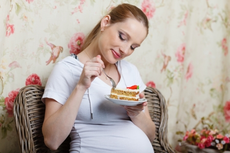 Young pregnant woman eating tasty cake sits on an armchair at homeの写真素材